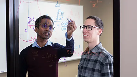 Two male students performing math equations on a glass wall.